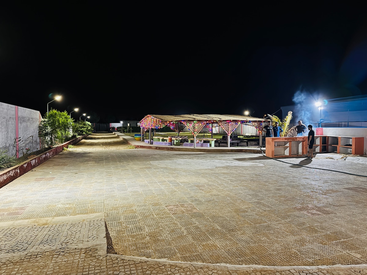 A well-lit pathway leads to a spacious outdoor area featuring a covered seating arrangement decorated with colorful lights. The surrounding greenery is subtly illuminated, and a barbecue setup is positioned nearby, contributing to a warm and inviting ambiance during the evening.