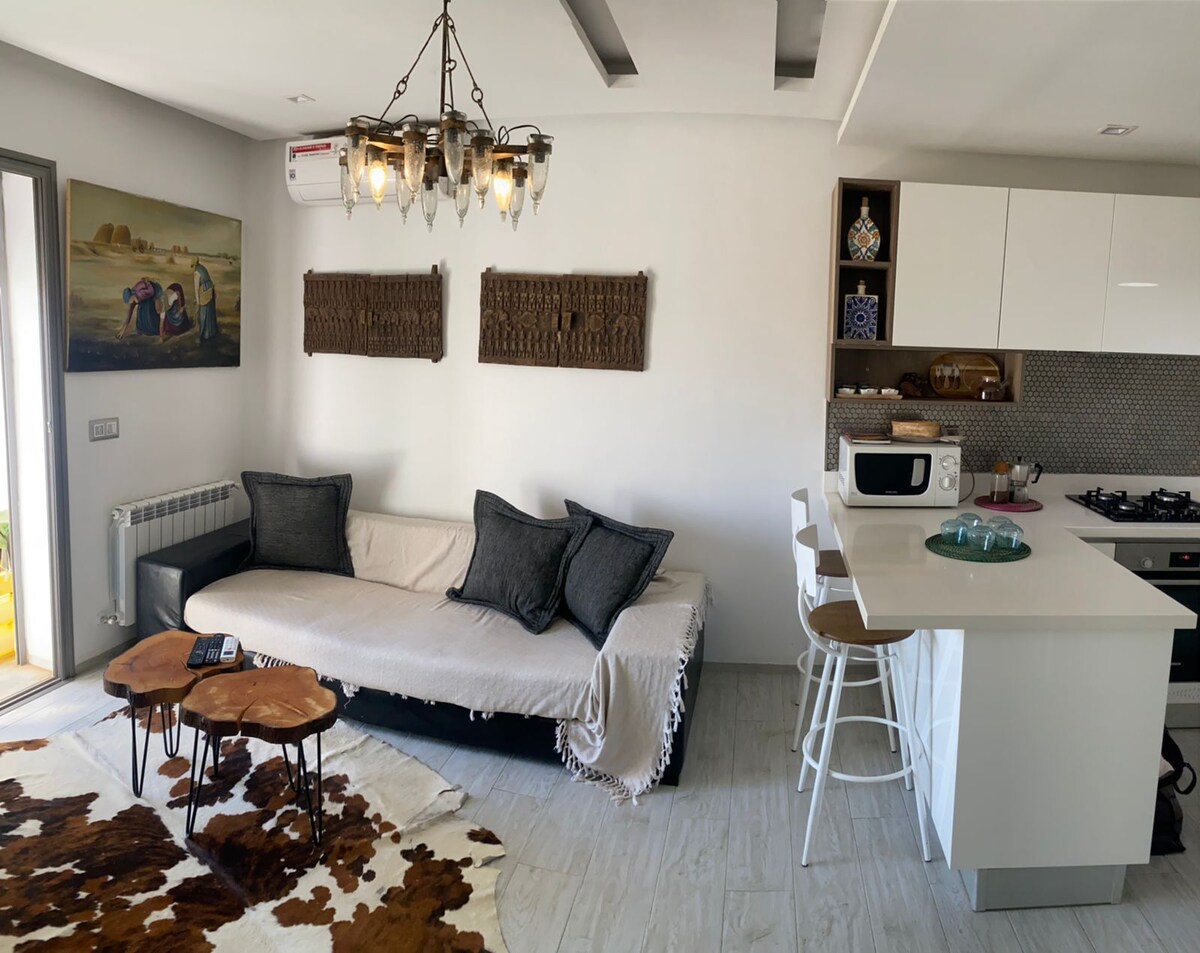 A modern living area features a comfortable sofa adorned with decorative pillows. A minimalist kitchen showcases sleek cabinetry, a microwave, and a gas stove. Unique artwork and wooden wall decor add character to the space. A patterned rug complements the light-colored flooring.