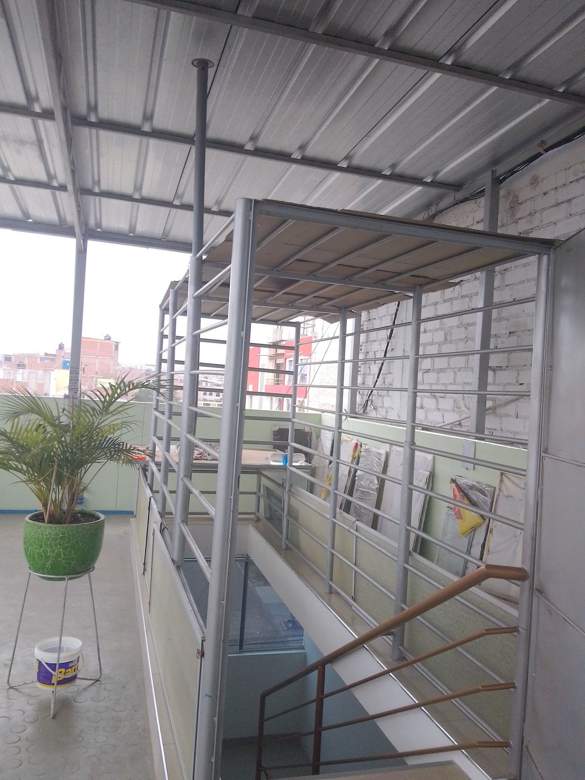 A spacious multi-level area shows a metal framework structure with an upper platform. A potted plant adds a touch of greenery, while the wide staircase leads to the lower level. Large windows provide natural light and views of the surrounding area.