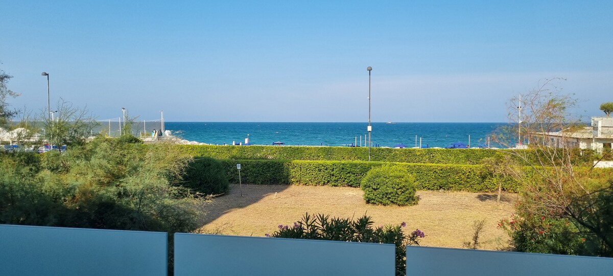 A view of the sea is presented, showcasing calm blue waters meeting a clear sky. The foreground includes a landscaped area with green shrubs and small flowering plants, while the distant horizon features gentle waves. Beach chairs are visible to the left, enhancing the coastal setting.