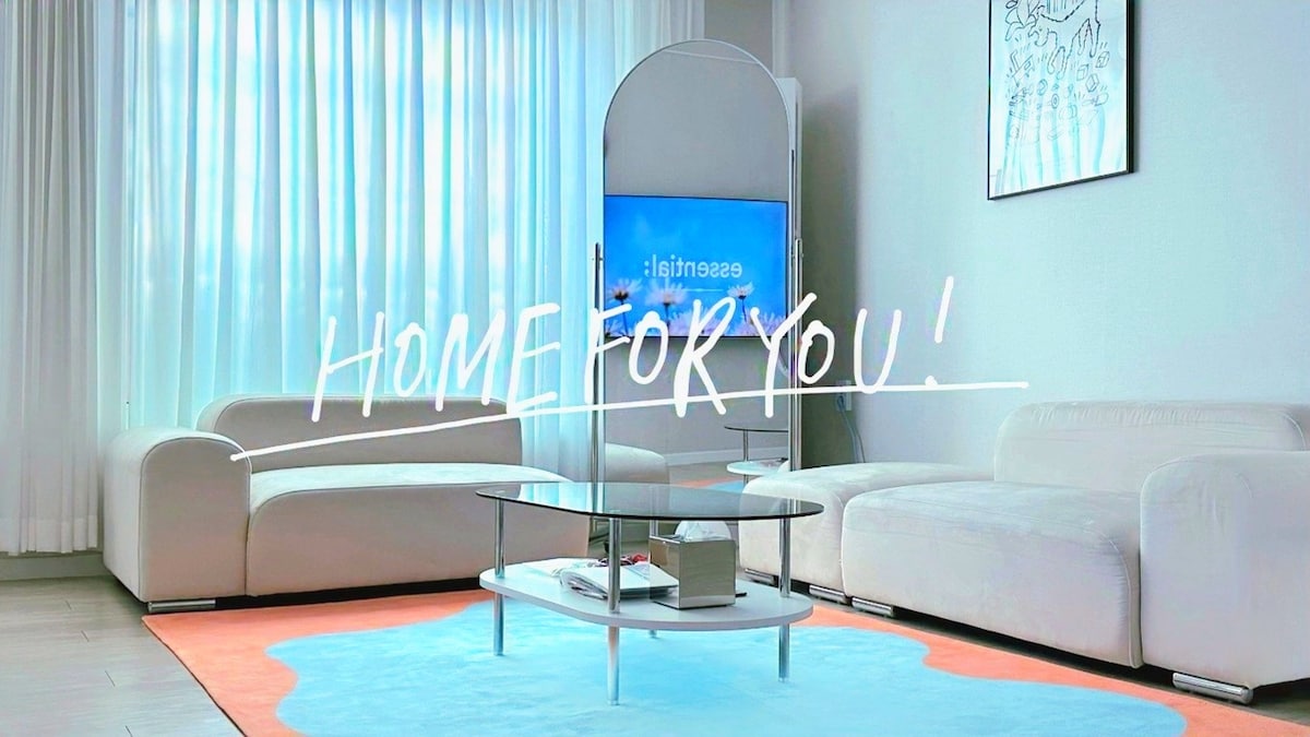 A spacious living area features two light-colored sofas arranged around a modern glass coffee table. A large smart TV is positioned across from the seating, and natural light filters through sheer curtains, illuminating the soft rug beneath.