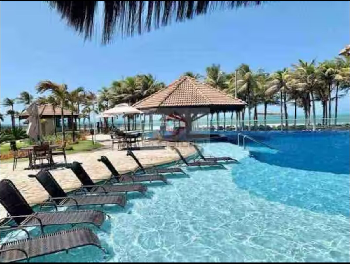 A serene pool area is featured, with a spacious infinity pool merging with ocean views. Sun loungers are arranged along the poolside, surrounded by palm trees. A shaded gazebo offers a relaxing spot, with umbrellas providing additional shade. The clear sky enhances the tranquil ambiance.