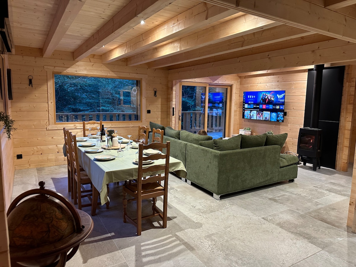 The spacious common area features a large dining table set for meals, adjacent to a comfortable seating arrangement. A wall-mounted screen displays entertainment options, while windows provide a view of the surrounding forest. The warm wood interior adds a cozy touch.