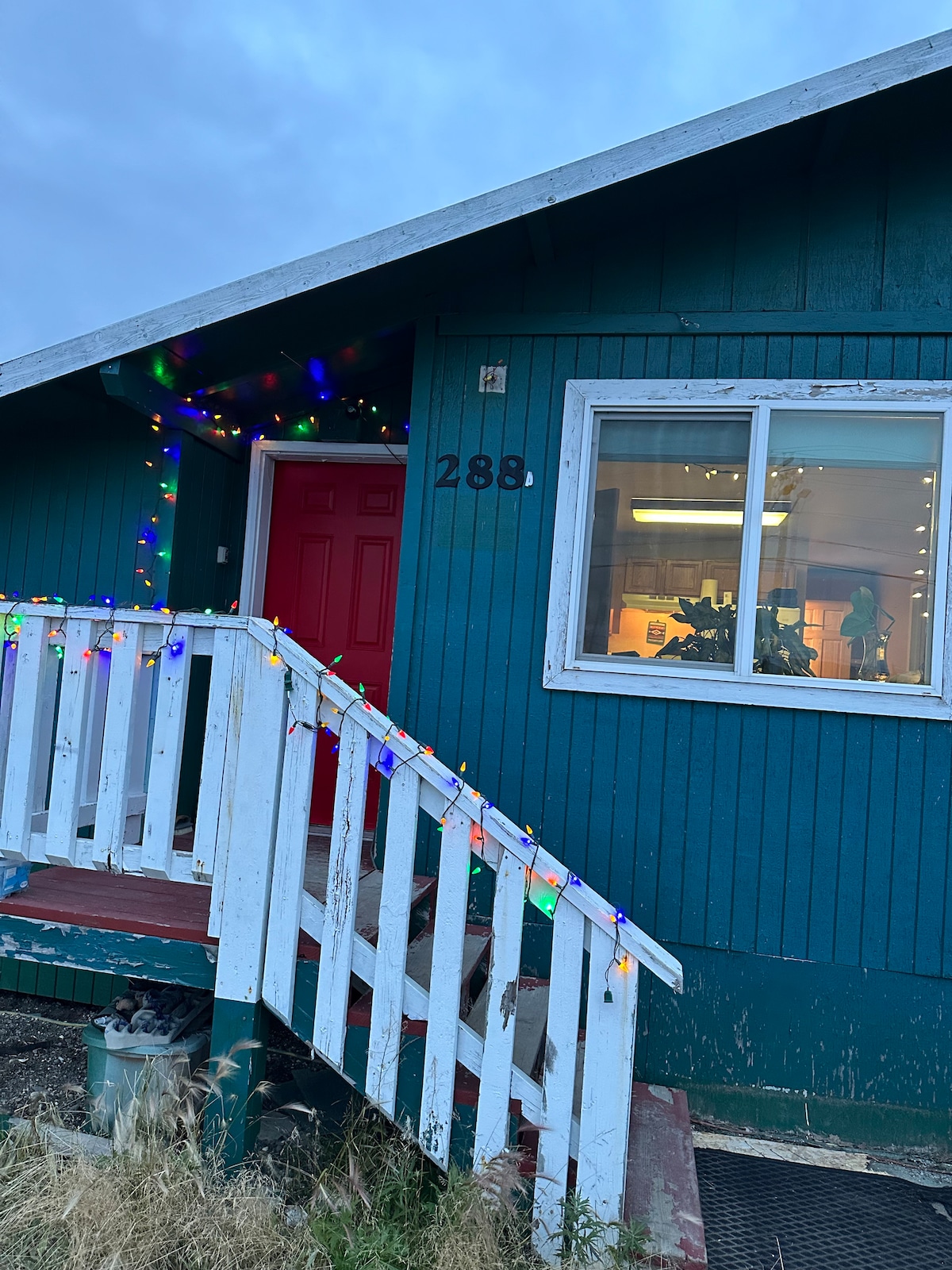 The exterior features a blue house with a welcoming red front door and decorative lights framing the entrance. A white railing leads up to the porch, providing a cozy touch. A large window reveals a softly lit interior, hinting at the warmth within.