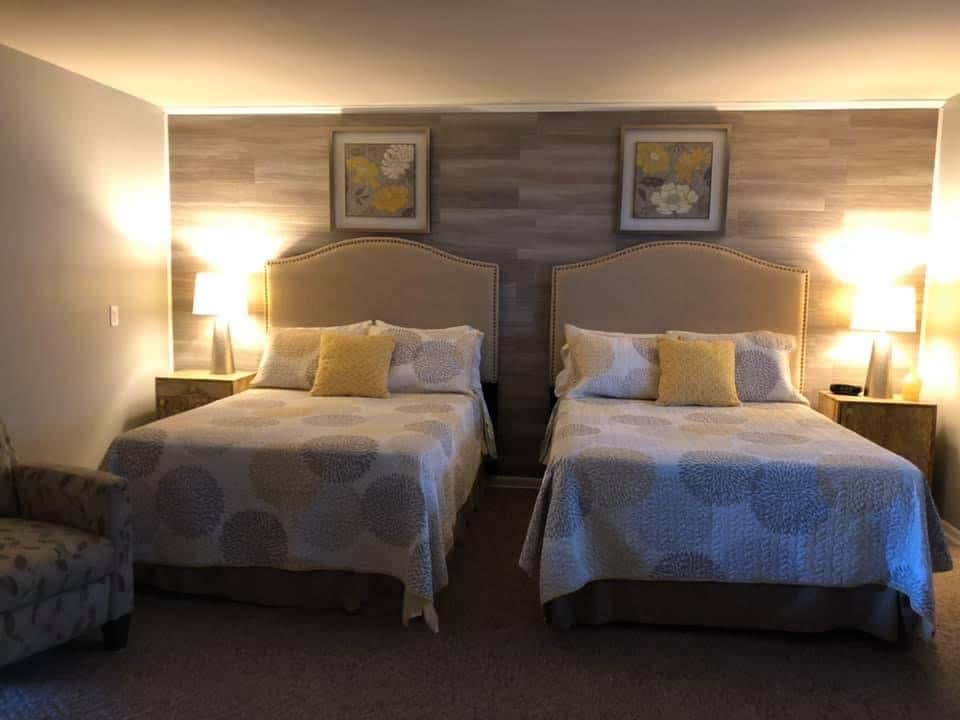 A comfortable sleeping area features two spacious beds, each adorned with light-colored bedding and decorative pillows. Nightstands flank the beds, providing lamps for soft illumination. A patterned chair is positioned nearby, and artwork hangs on the wall above the beds, adding subtle decor.