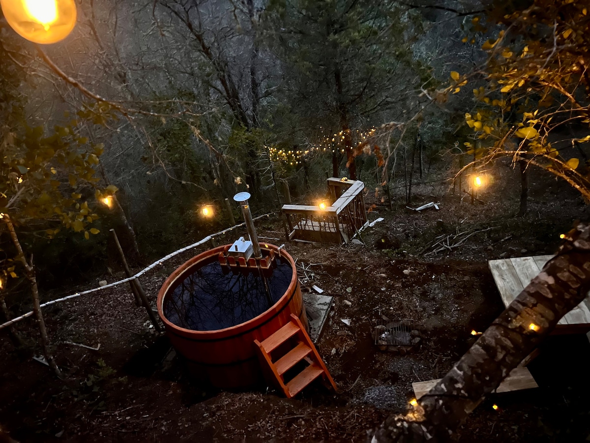 A wooden hot tub is positioned among trees, surrounded by soft outdoor lighting. Steps lead into the tub, while a wooden platform is visible nearby, providing a relaxing space to unwind in nature.