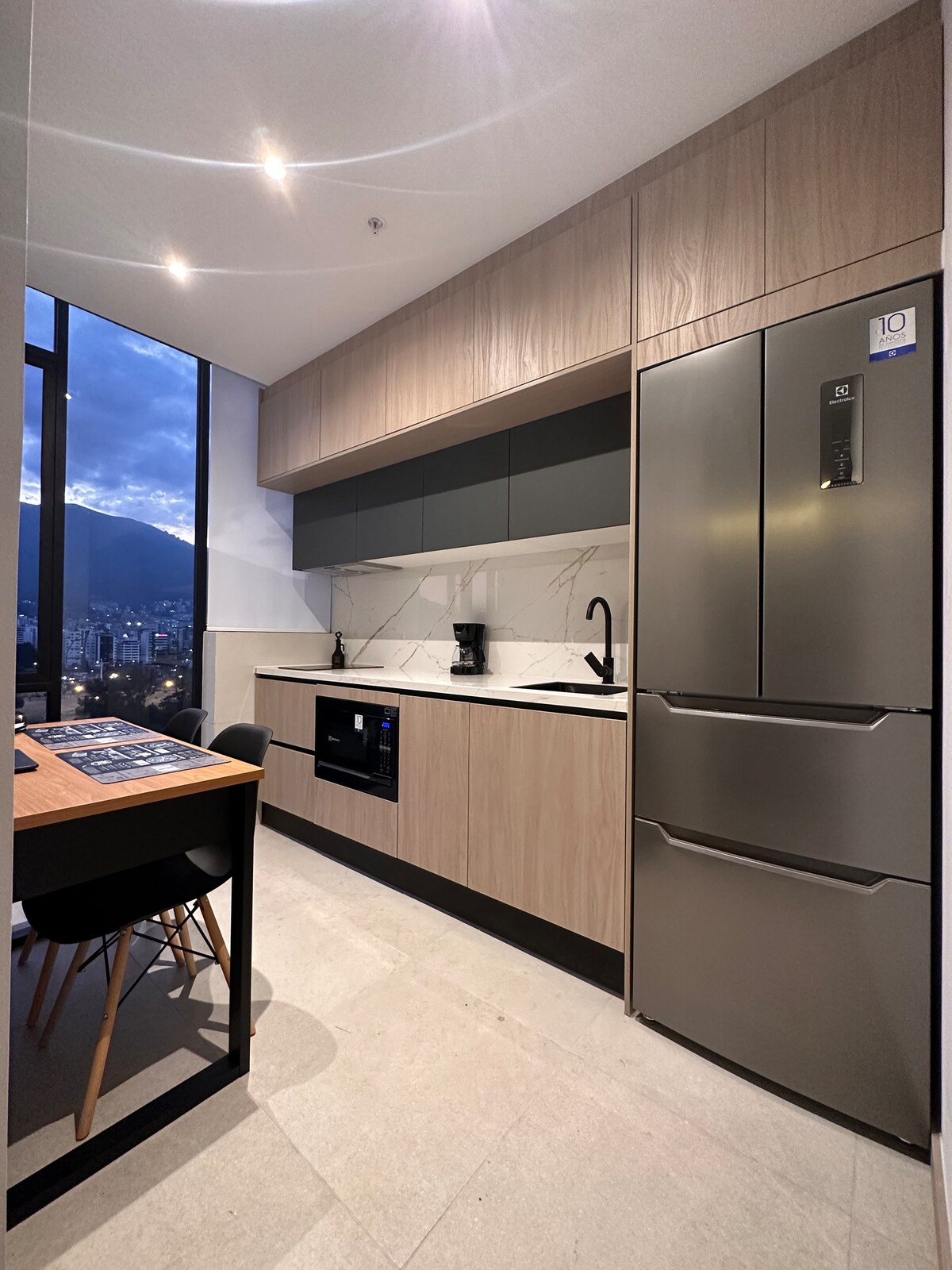A modern kitchen area is presented with sleek cabinetry in light and dark tones. A countertop with a sink and stove is visible, complemented by a built-in microwave and stainless steel refrigerator. A small dining table with black chairs is positioned near the expansive window showing city views.