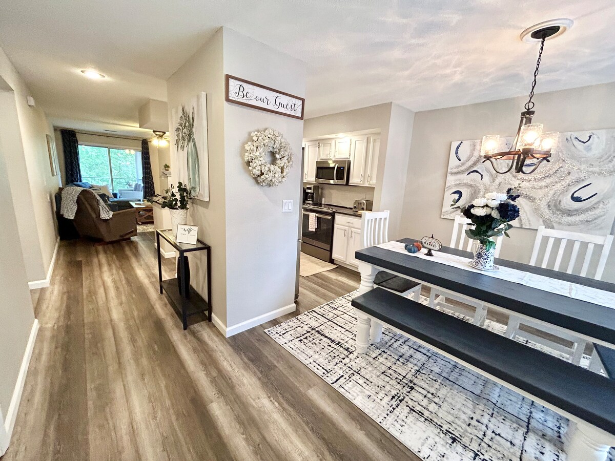 A spacious dining area features a long dark dining table with white chairs. A chandelier hangs above, illuminating a floral centerpiece. Light wood flooring extends into an adjoining open kitchen, which is visible in the background along with a cozy living area featuring a sofa.