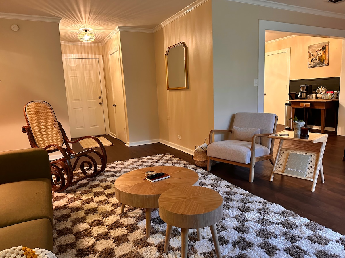 A welcoming living area features a comfortable rocking chair and a light-colored armchair. Two round wooden tables are positioned on a textured area rug. A mirror reflects the soft lighting, while the entrance and kitchen areas are visible in the background.