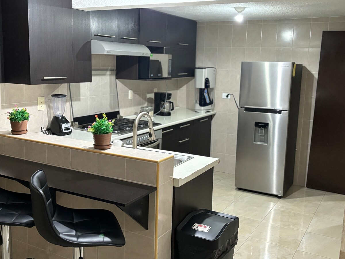 A modern kitchen is equipped with dark cabinetry and stainless steel appliances, including a refrigerator and stove. A breakfast bar with two black stools is positioned adjacent to the kitchen counter, which features a small potted plant for a touch of greenery.