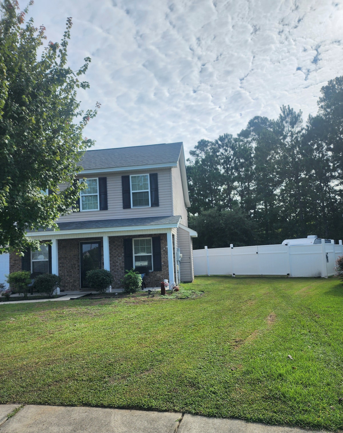 A two-story building is shown with a neatly maintained lawn in front. The façade features dark shutters and a welcoming entrance. A fenced yard is visible in the background, complemented by trees that create a natural backdrop.