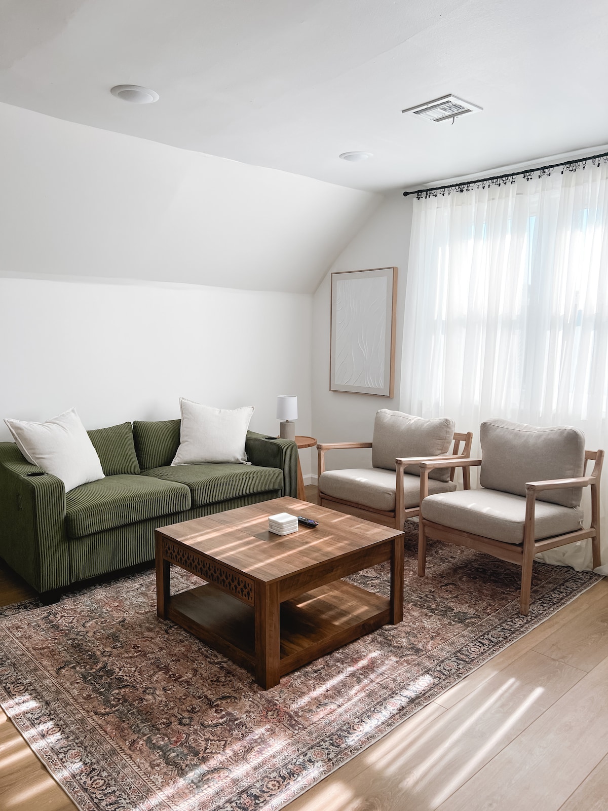 A bright living area is presented with a green sofa and two light-colored lounge chairs positioned around a wooden coffee table. Natural light streams through the sheer curtains, illuminating the space adorned with a decorative area rug.
