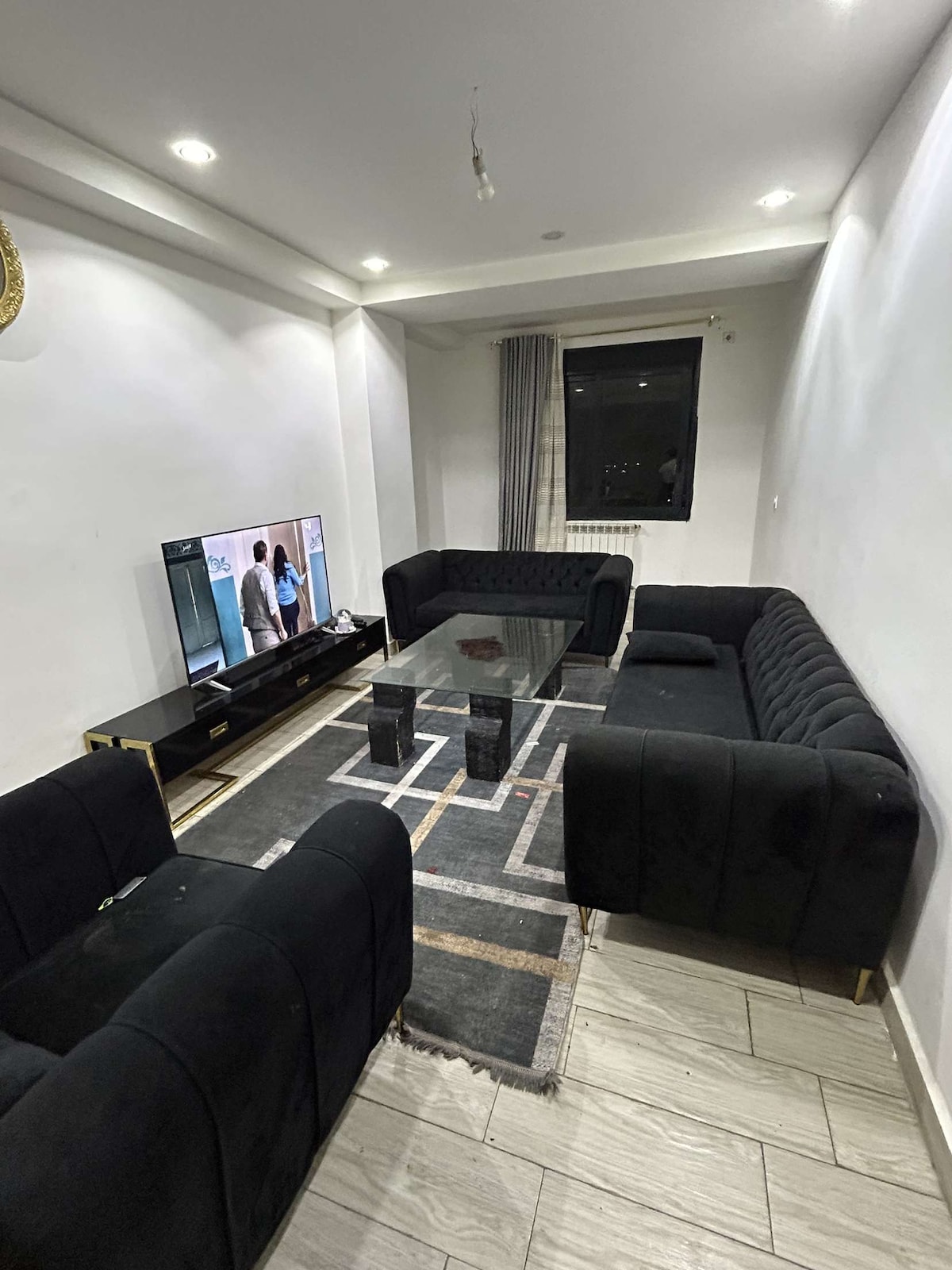 A modern living area features a sleek black sofa set arranged around a rectangular glass coffee table. A television is positioned against the wall, reflecting soft lighting. Light gray curtains frame a window, adding a subtle contrast to the room's design.