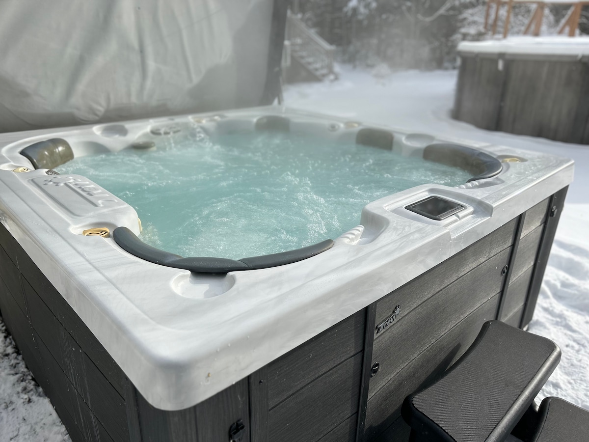 A spacious hot tub sits surrounded by a snowy landscape, with steam rising gently from the warm water. The tub features comfortable seating and an integrated control panel. Nearby, a soft step provides easy access for guests, enhancing a relaxing outdoor experience.