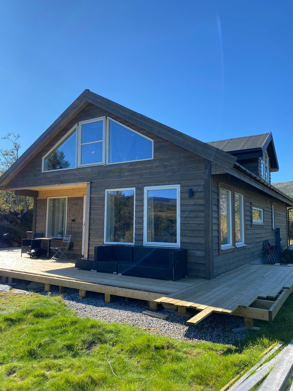 A wooden cabin features large windows and a sloped roof, surrounded by green grass. A spacious deck extends from the front, accommodating outdoor seating. Natural lighting enhances the inviting facade, which blends seamlessly with the surrounding landscape.