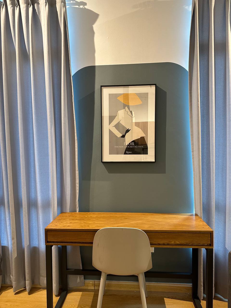 A compact workspace is presented with a wooden desk and a white chair, positioned against a wall featuring a geometric design. The wall is painted in two colors, with a framed art piece mounted above the desk. Light grey curtains frame the window, offering a muted ambiance.