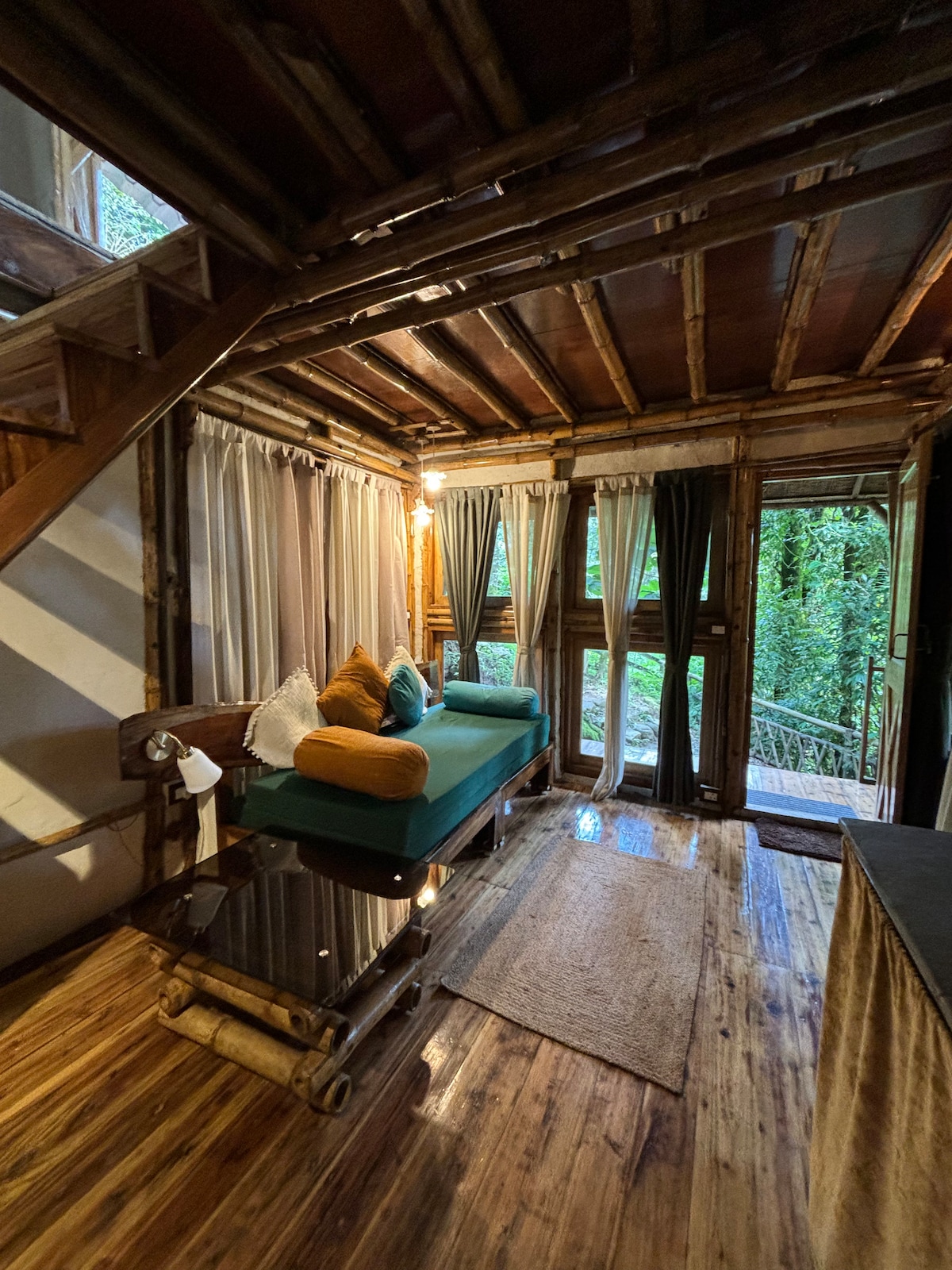 A cozy living area features a green sofa adorned with decorative pillows, placed beside a wooden table. Natural light enters through large windows dressed in light curtains, illuminating the wooden walls and flooring. A woven rug adds texture to the space.