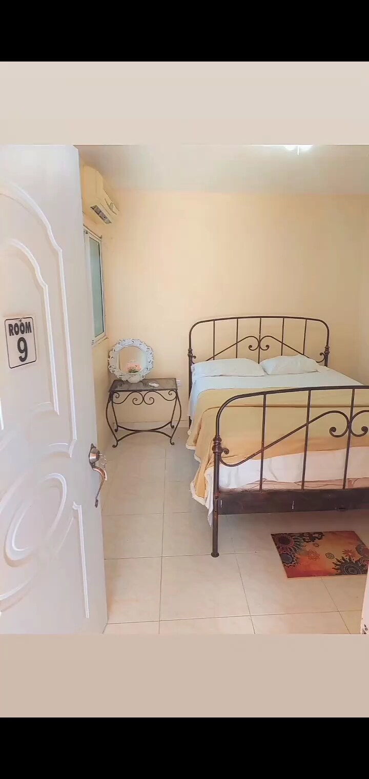 A small bedroom features a metal bed with a light yellow blanket, complemented by a simple nightstand. Natural light filters through a window, illuminating the soft-colored walls. A decorative mirror is positioned on the wall above the nightstand, adding a functional element to the space.