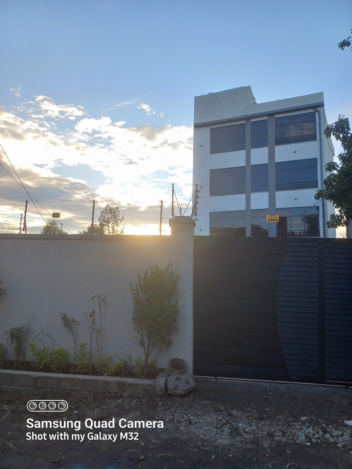 The exterior of a modern building is seen against a backdrop of a sunset, creating a serene atmosphere. A high wall with a black gate provides privacy, while small shrubs are planted along the base, enhancing the landscaped area in front.