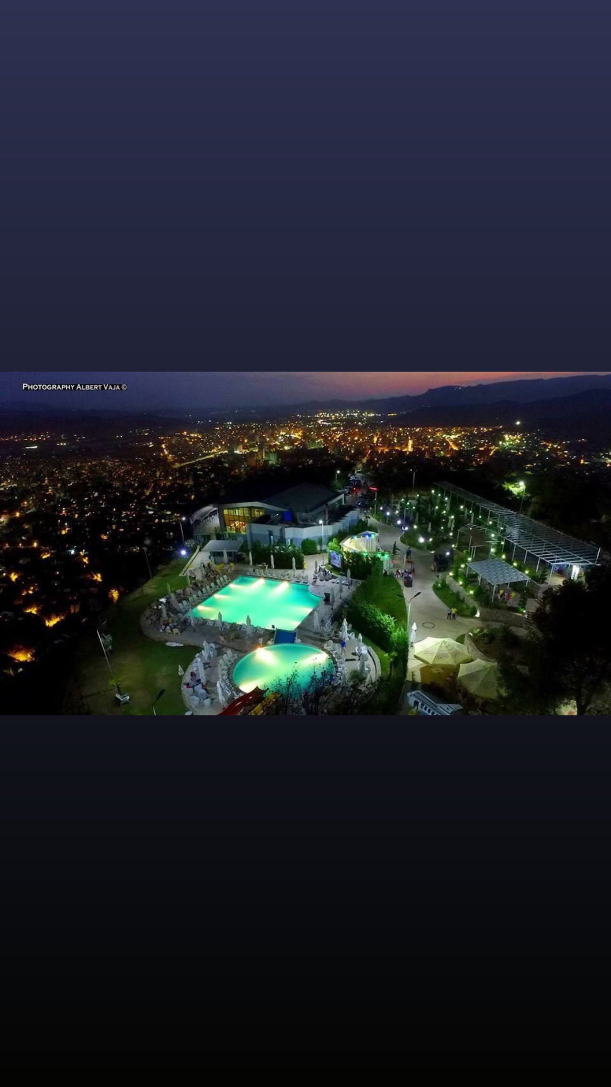 An aerial view captures a vibrant outdoor area illuminated by soft lighting. A swimming pool is visible, surrounded by lush greenery and seating arrangements, with multiple terraces and pathways. A picturesque hillside community is set in the background, showcasing twinkling city lights at dusk.