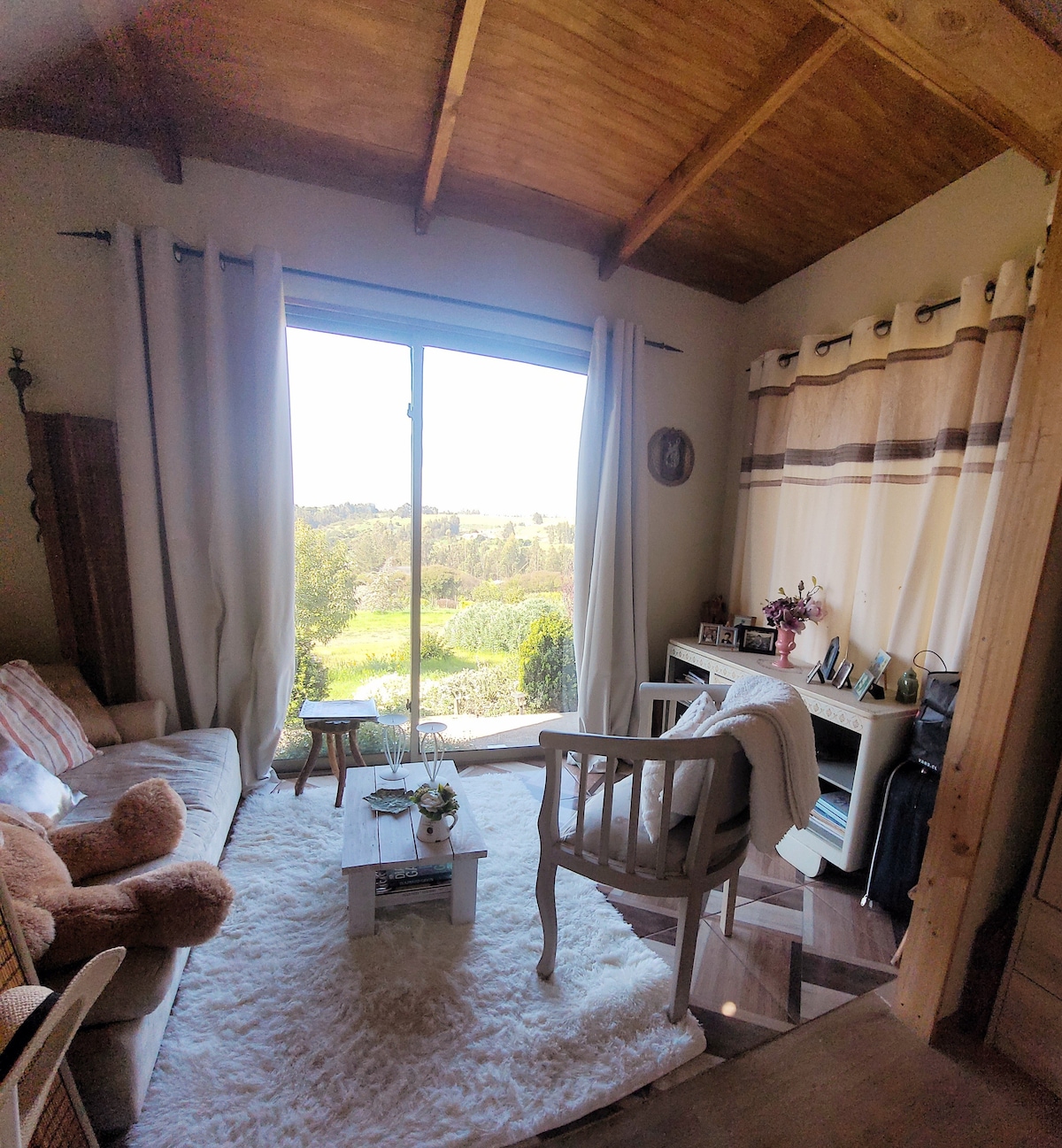 A cozy sitting area features a light-colored sofa and a white armchair, oriented towards a large glass door that offers views of the lush greenery outside. A small coffee table is adorned with decorative items, and natural light fills the space through the curtains.