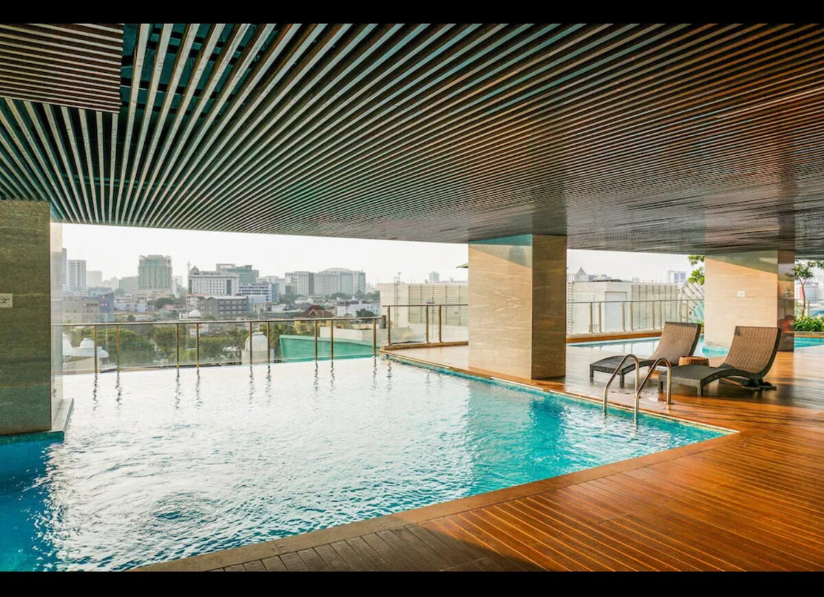 An outdoor swimming pool is featured on a high floor, surrounded by sleek wooden decking. Loungers are positioned along the edge, offering views of the city skyline through expansive glass panels. The pool's clear water reflects the modern architectural elements overhead.
