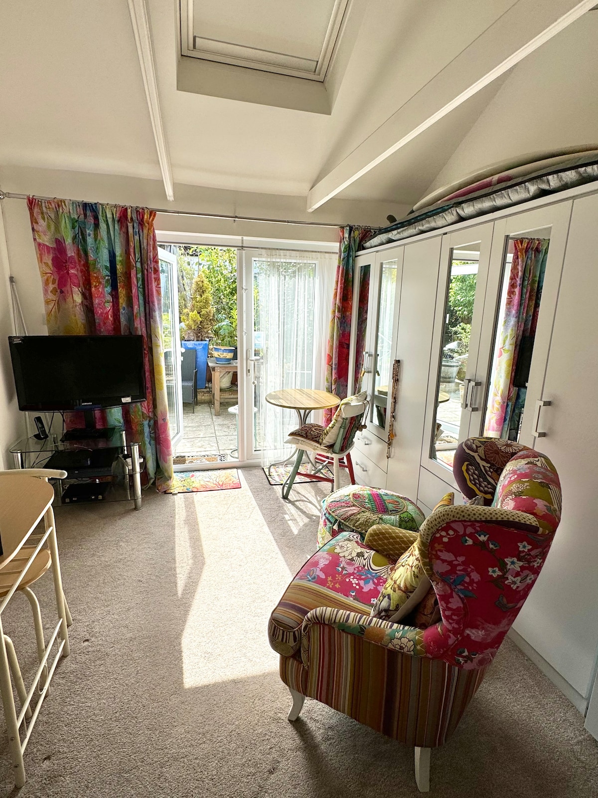 A bright living area features a colorful floral-patterned armchair and a small round table. Large glass doors lead to the patio, allowing natural light to fill the space. Curtains with vibrant designs frame the door, complementing the cheerful decor.