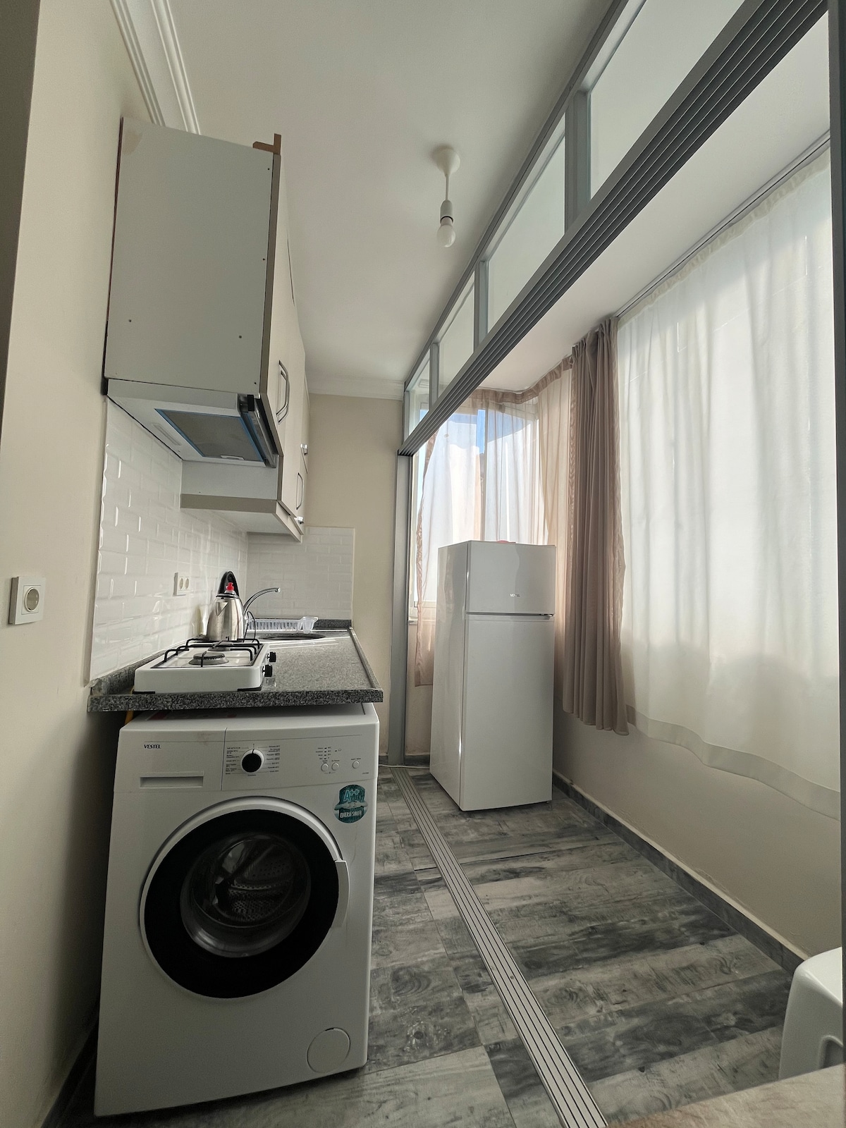 A compact kitchen area features modern appliances, including a washing machine and a refrigerator. Natural light floods through large windows lined with light curtains, illuminating the stone countertop and sleek cabinetry. The floor displays stylish tiles, providing a neat and functional space.