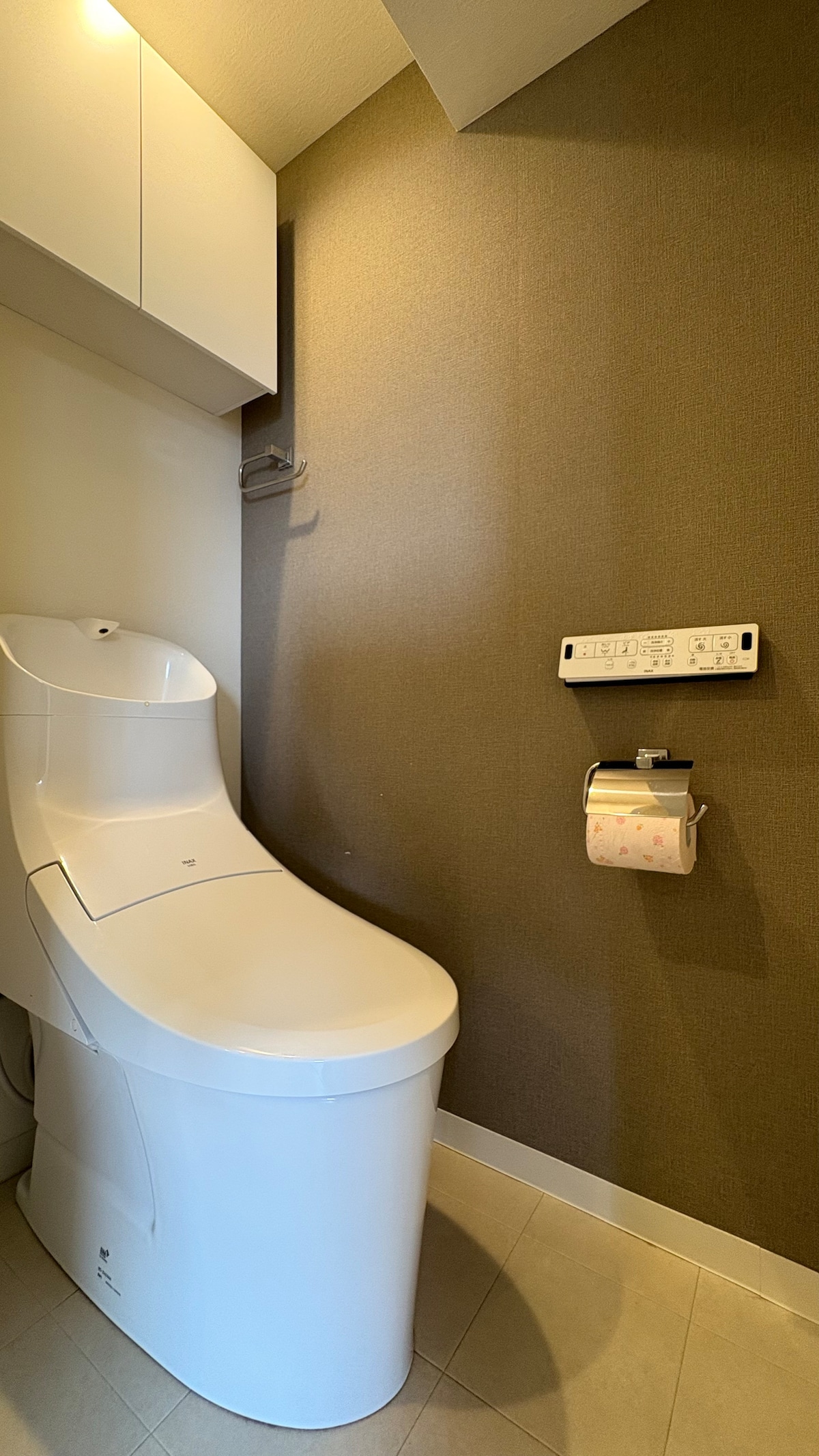 A modern bathroom is displayed featuring a sleek toilet design against a textured wall. A roll of toilet paper is mounted nearby, with additional storage options seen above, providing a functional and tidy space.