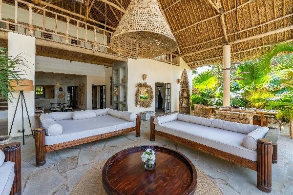 An open living area features high thatched ceilings and natural stone flooring. Two spacious, low-sitting sofas with neutral cushions are positioned around a round wooden coffee table. Plants are visible in the background, contributing to a serene atmosphere.