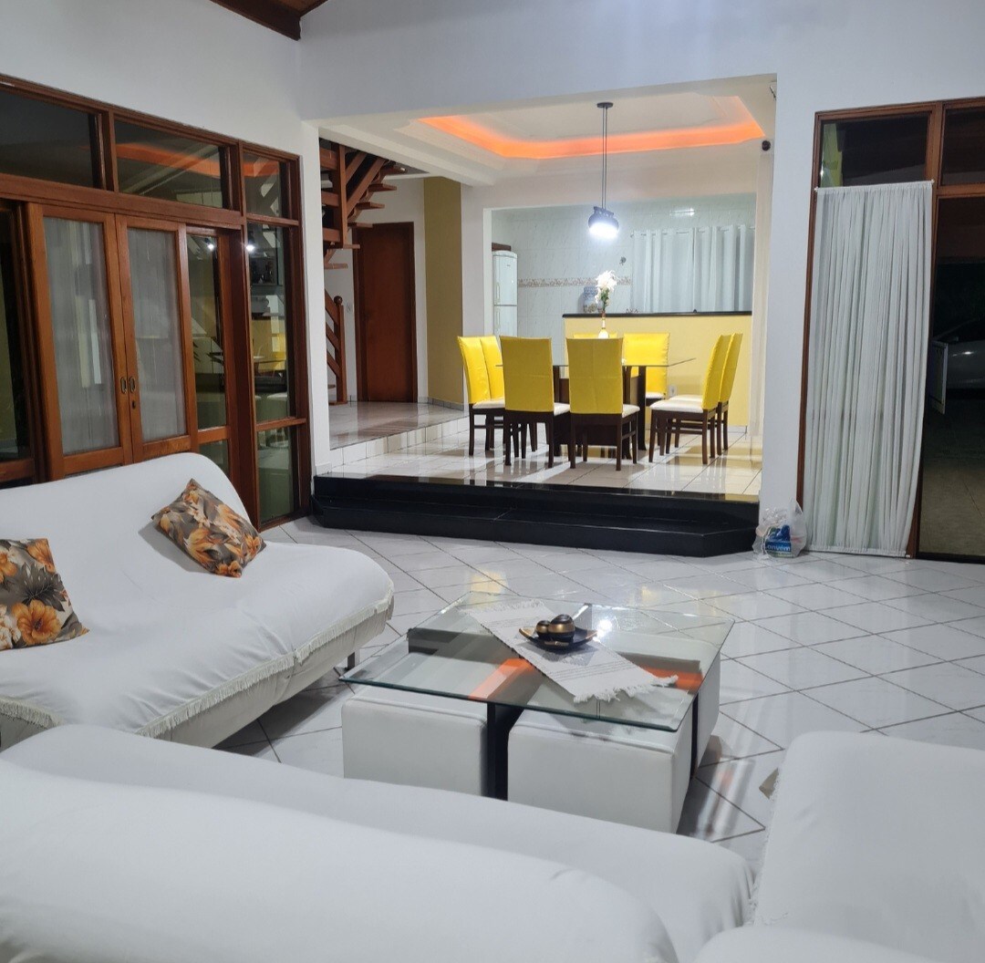 A spacious living area is furnished with white sofas and decorative cushions. A glass coffee table is centered among the seating. The dining space in the background features a large table surrounded by yellow chairs, with a kitchen visible beyond.