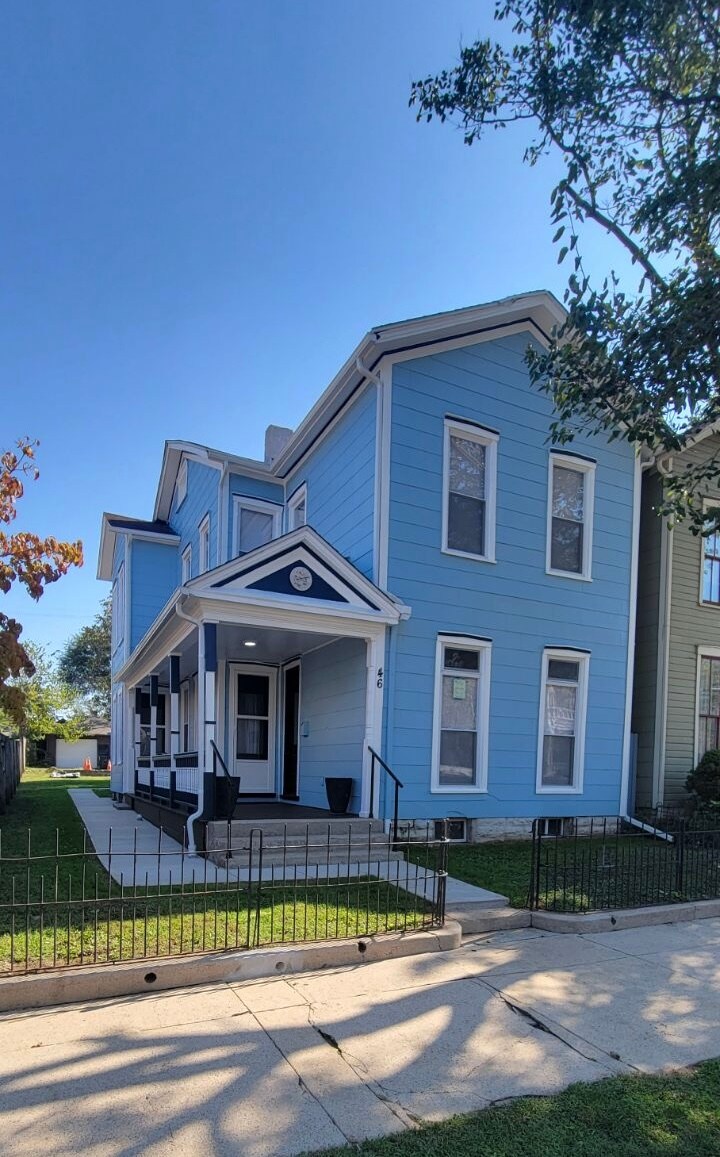 A light blue two-story home is presented, featuring a neatly trimmed lawn and sidewalk. The entrance is framed by steps leading to a covered porch, with large windows that provide ample natural light. The fence surrounding the property offers privacy and a welcoming appearance.