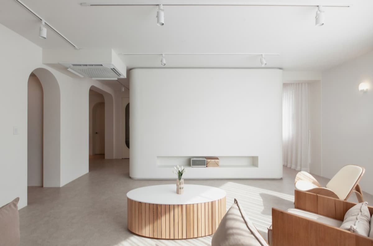 A modern living space features a large circular coffee table made of wood, complemented by a light-colored armchair and sofa. The room is illuminated by track lighting, with soft natural light filtering through sheer white curtains. The minimalist design is highlighted by neutral tones and clean lines.