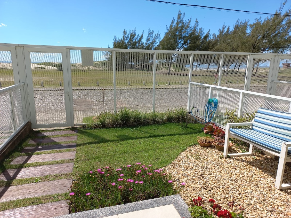 A well-manicured outdoor area is showcased, featuring a path of wooden slats leading to a grassy space. Colorful flowers add vibrancy, while a blue and white garden chair invites relaxation. A protective fence provides safety, with clear views of the nearby beach beyond.