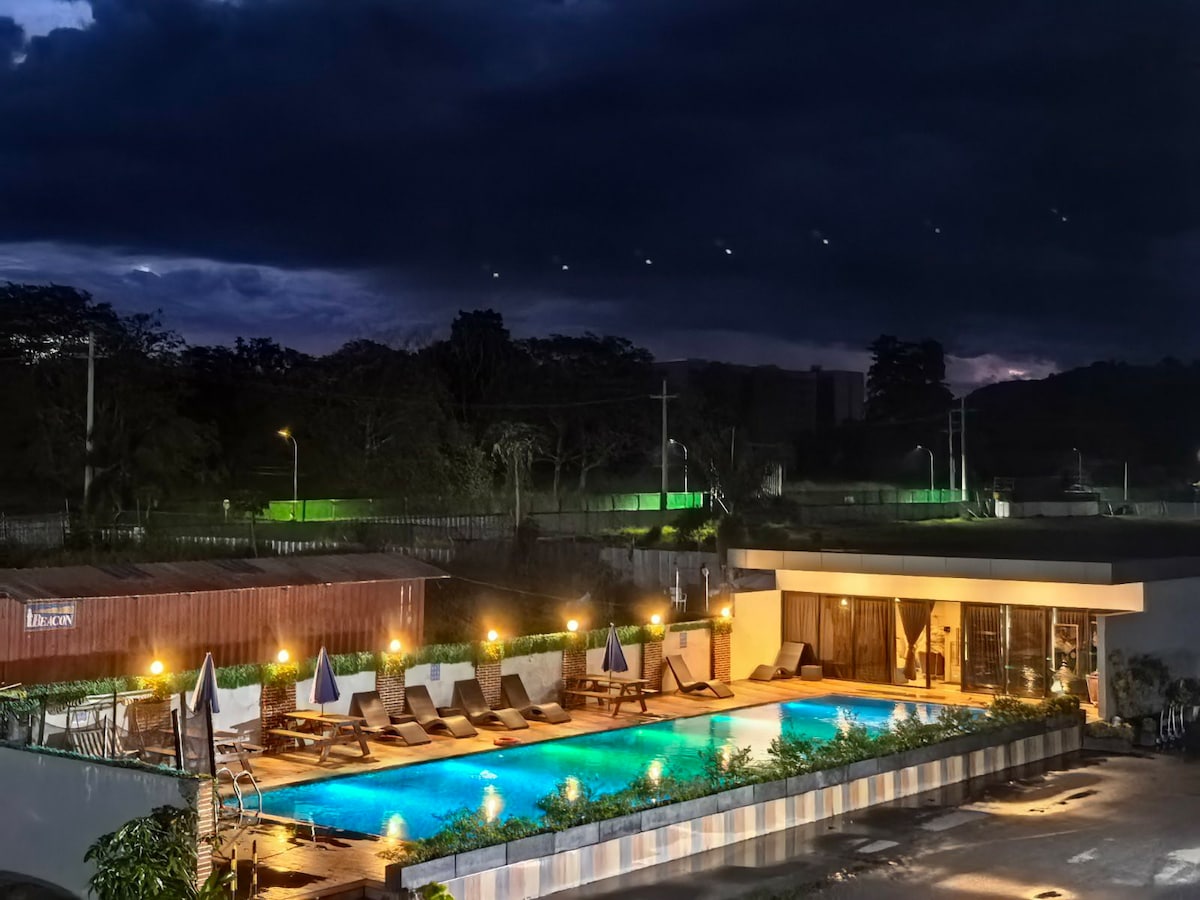 An outdoor swimming pool is surrounded by lounge chairs and umbrellas, inviting relaxation. Soft lighting enhances the atmosphere, while the building in the background features large windows. Dark clouds provide a dramatic backdrop, indicating an evening setting.