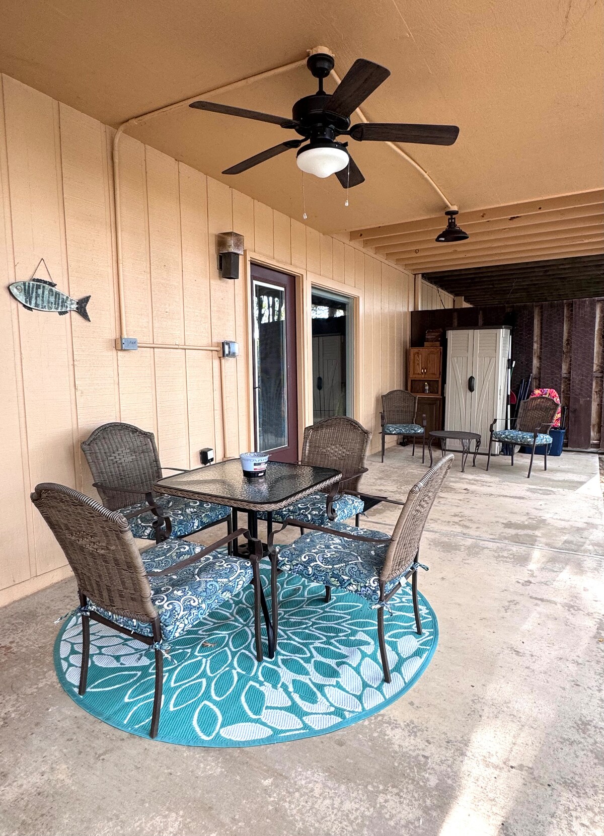 A covered patio features a round table surrounded by four chairs, all set on a circular, patterned rug. A ceiling fan hangs above, providing airflow. A storage unit is visible in the background, with additional seating options along the side.
