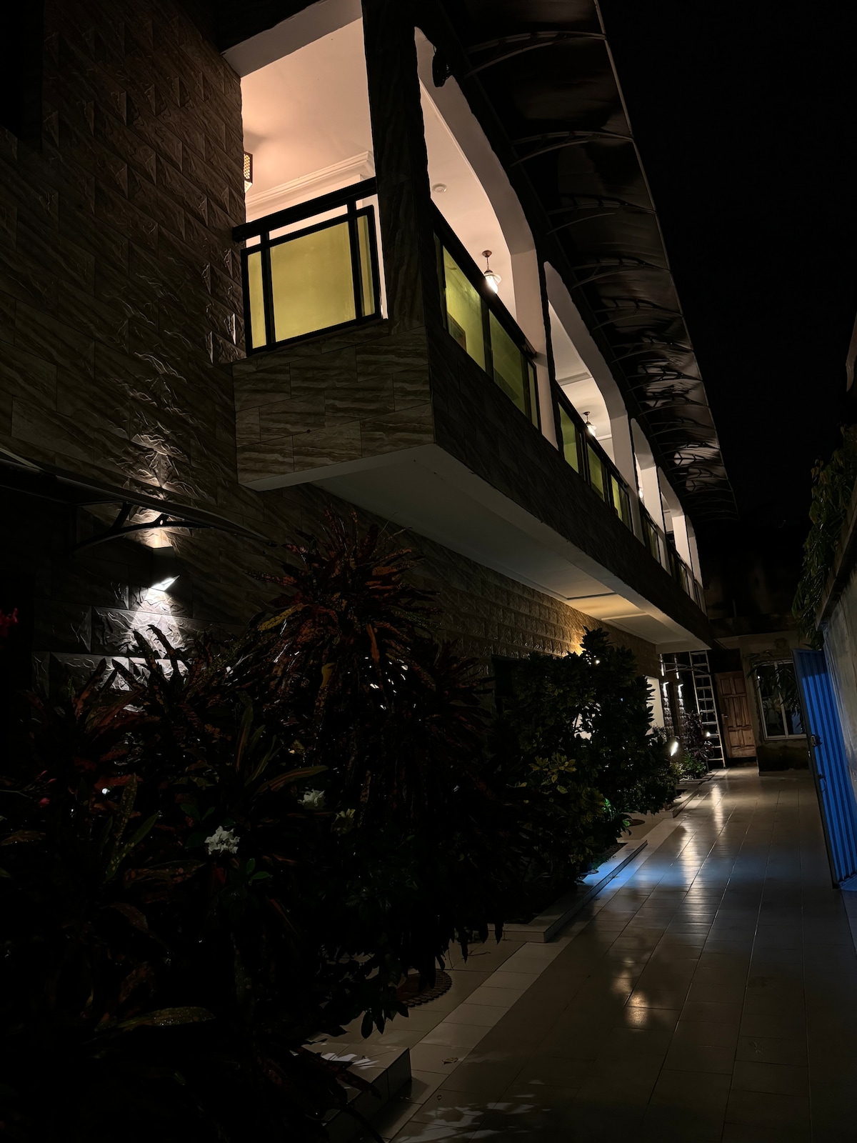 A pathway is bordered by lush greenery and well-lit walls, creating an inviting atmosphere. The upper level features a balcony with modern railings. Soft lighting enhances the texture of the stone exterior, offering a warm contrast to the night sky.