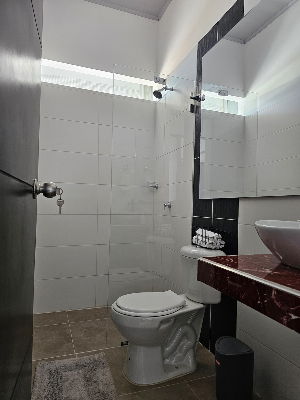 A modern bathroom is showcased, featuring a glass-enclosed shower with a rainfall showerhead. A white toilet and a sleek sink with a marble countertop are visible. Natural light enters through a frosted window, enhancing the bright, clean aesthetic.