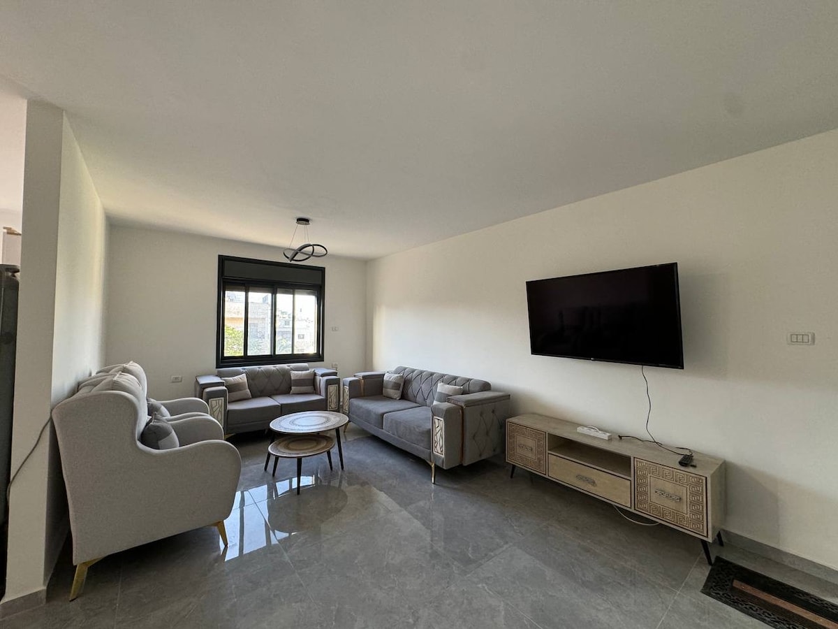 An open living area features a comfortable gray sectional sofa and two armchairs, arranged around a circular wooden coffee table. A mounted flat-screen television is visible on the wall, while natural light enters through a large window, adding to the spacious feel.