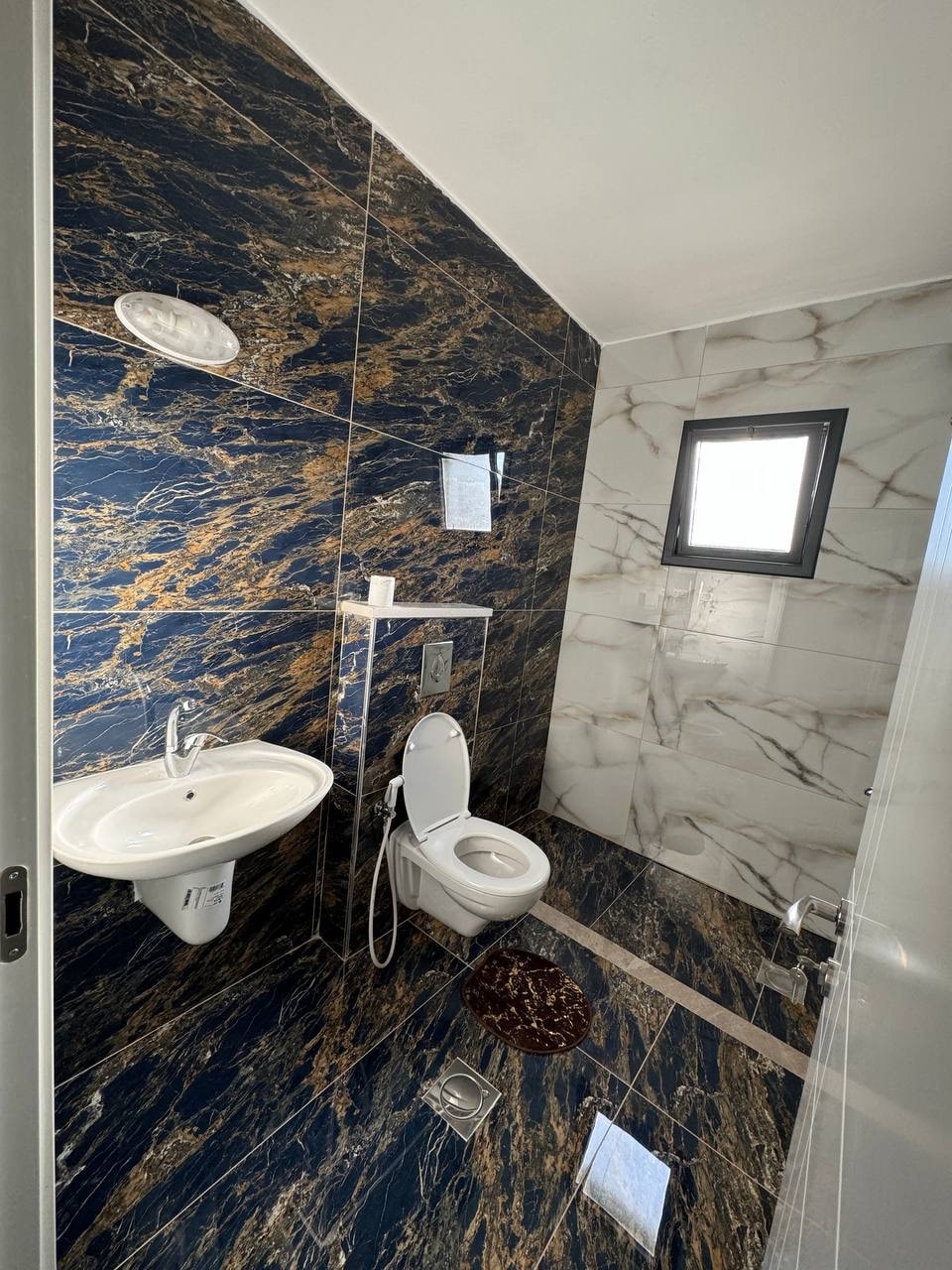 A modern bathroom is presented with elegant marble walls in blue and gold tones. A sleek, wall-mounted sink and a contemporary toilet are positioned against the stylish backdrop. Natural light enters through a small square window above, enhancing the bright and clean aesthetic.