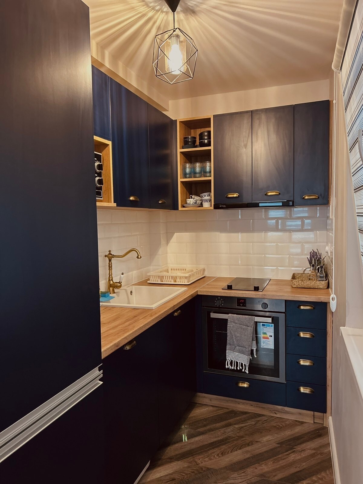 A compact kitchen is showcased with dark cabinetry and wooden countertops. Shelves above display glassware and kitchen essentials. A sink is situated under a window, while an oven and stovetop are integrated within the cabinetry. A pendant light adds illumination to the cozy space.
