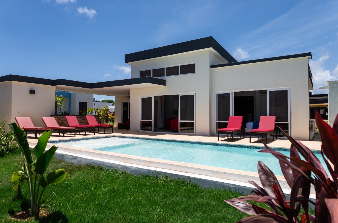 A modern villa is depicted featuring large glass windows and a patio area. The inviting private pool is surrounded by well-maintained grass and several red lounge chairs. Lush plants are visible in the foreground, contributing to the tropical setting.