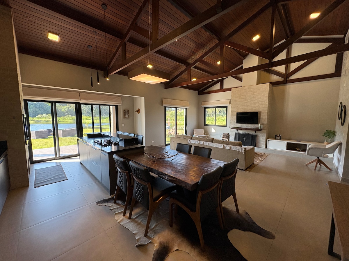 An open-concept living and dining area is depicted, featuring a large wooden dining table with comfortable chairs. Expansive windows allow natural light to flood the space, showcasing views of the surrounding landscape. A modern kitchen with sleek cabinetry is visible, and a cozy seating area is arranged nearby.