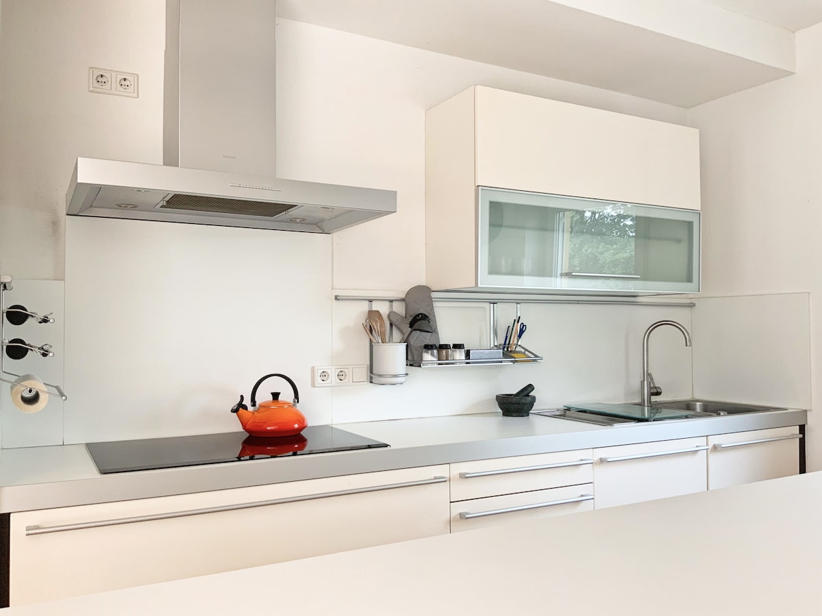 The kitchen features modern white cabinetry and a sleek countertop. An orange kettle is placed on the stove, alongside cooking utensils stored on the wall. A sink is located to the right, with natural light illuminating the space through a nearby window.