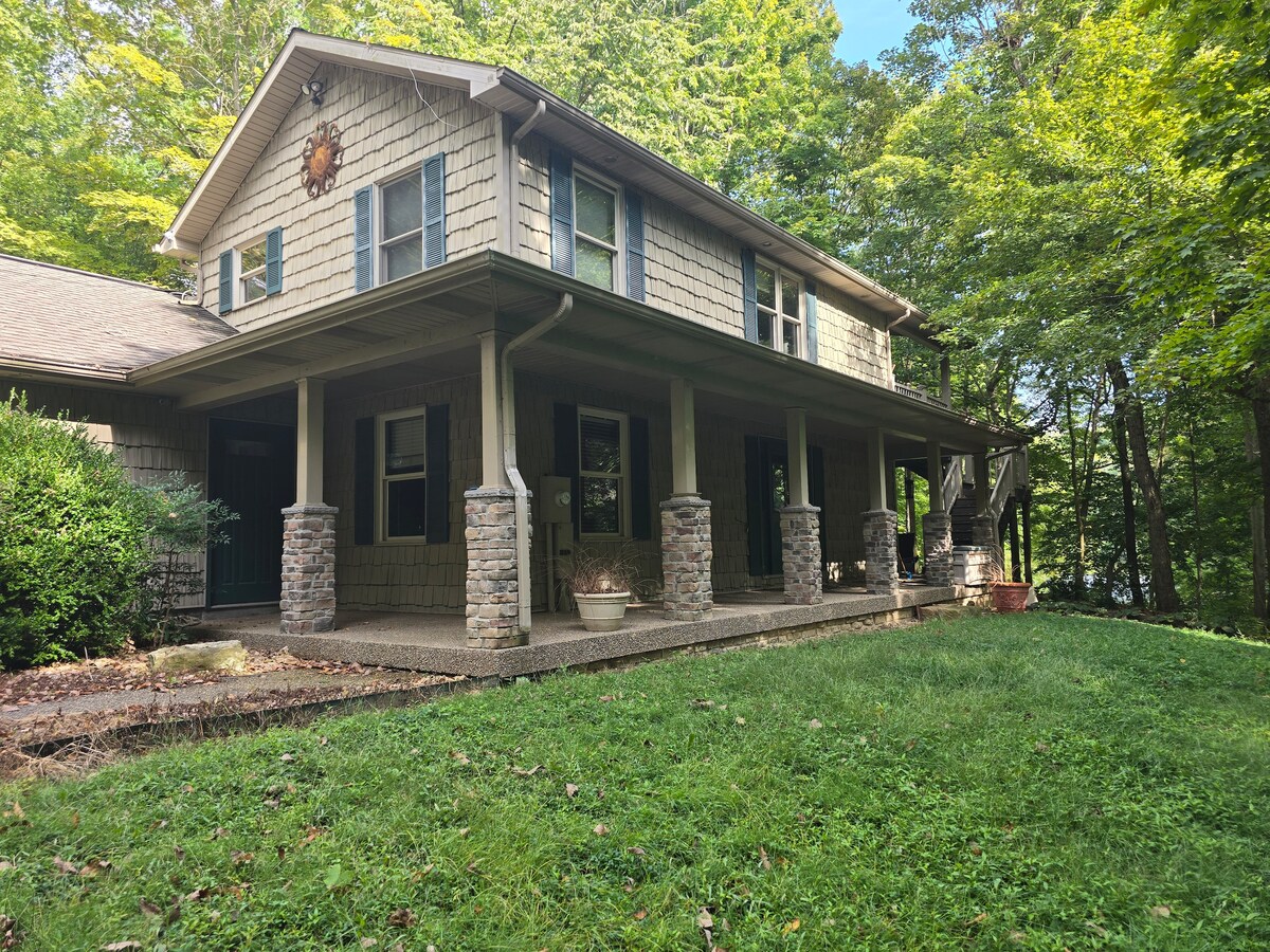 The exterior of a two-story cabin is displayed, featuring a covered porch supported by stone columns. Lush greenery surrounds the property, with a manicured lawn and trees providing shade, creating a serene and natural setting.
