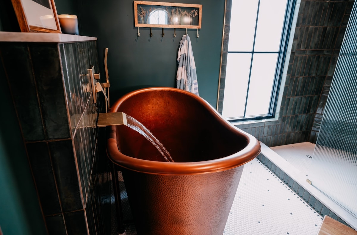 A deep copper soaking tub is positioned against a dark green wall, featuring a wooden spout for water flow. Natural light illuminates the space through a large window, while a stylish shower area and sleek, patterned tiles complement the luxurious ambiance.