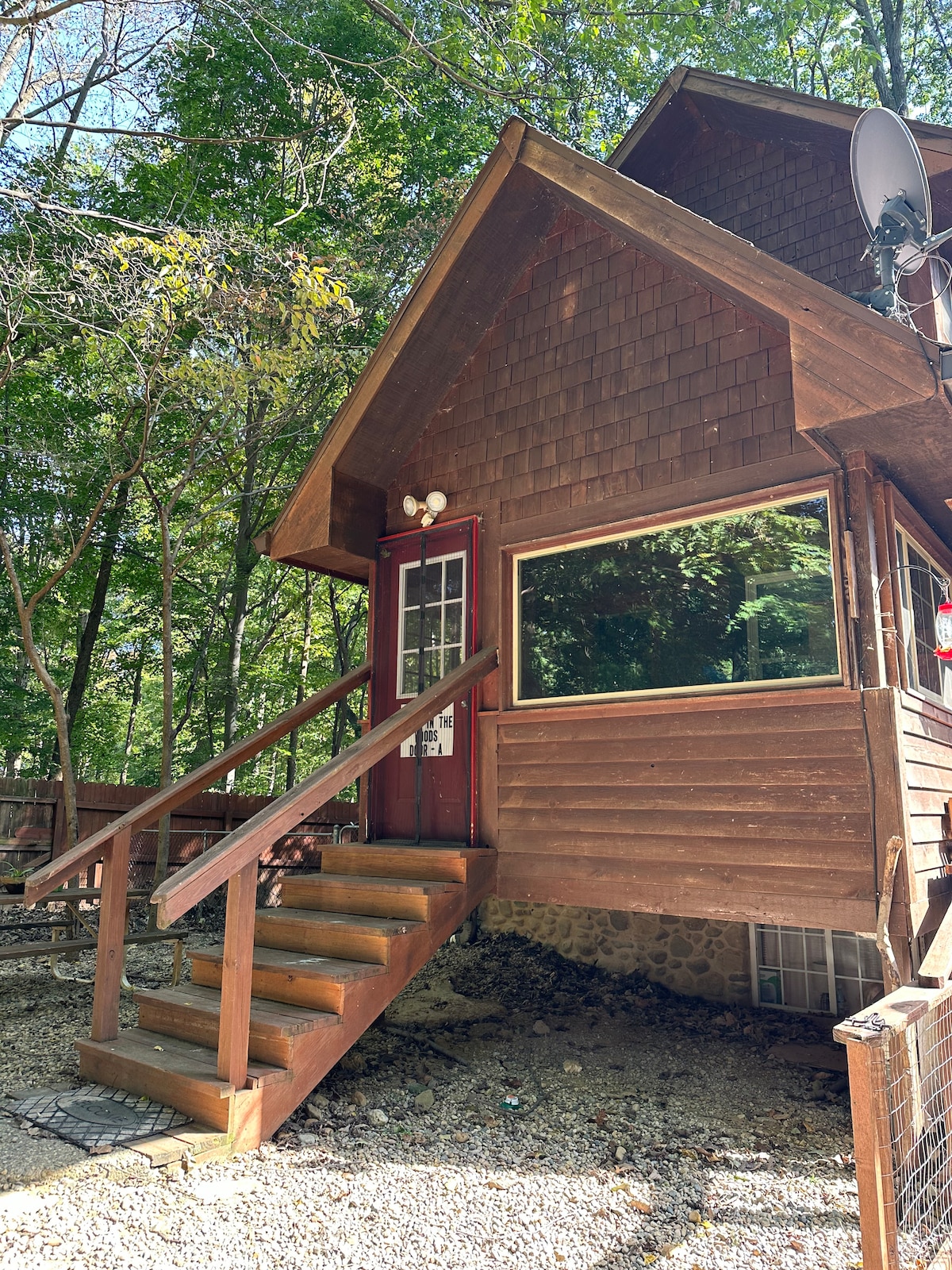 The entrance features a set of wooden steps leading up to a rustic cabin with a shingled roof. A large window offers views of the surrounding greenery, while a satellite dish is mounted to the side of the building.