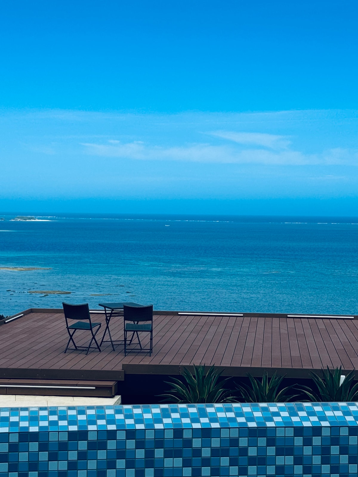 A wooden deck overlooking the ocean features two chairs and a small table, providing a serene spot for relaxation. The vibrant blue waters stretch out under a clear sky, creating an inviting atmosphere for enjoying the coastal views.