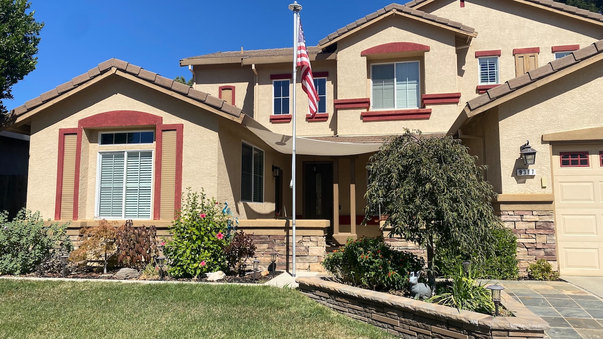The exterior of a spacious house is displayed, featuring a welcoming porch and colorful shrubs. An American flag is proudly flown, while the stone accents provide character. The bright blue sky creates a lively backdrop, enhancing the curb appeal of the home.