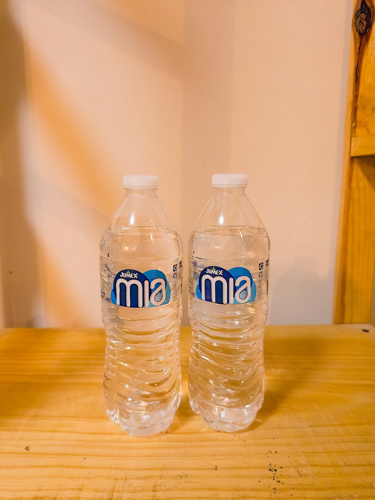 Two clear plastic water bottles are displayed on a wooden surface. Each bottle is filled with water and features a label with the brand 'mia.' Natural light is softly illuminating the scene.